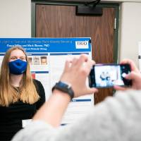 Guest taking photo of scholar and mentor in front of poster, all wearing masks.